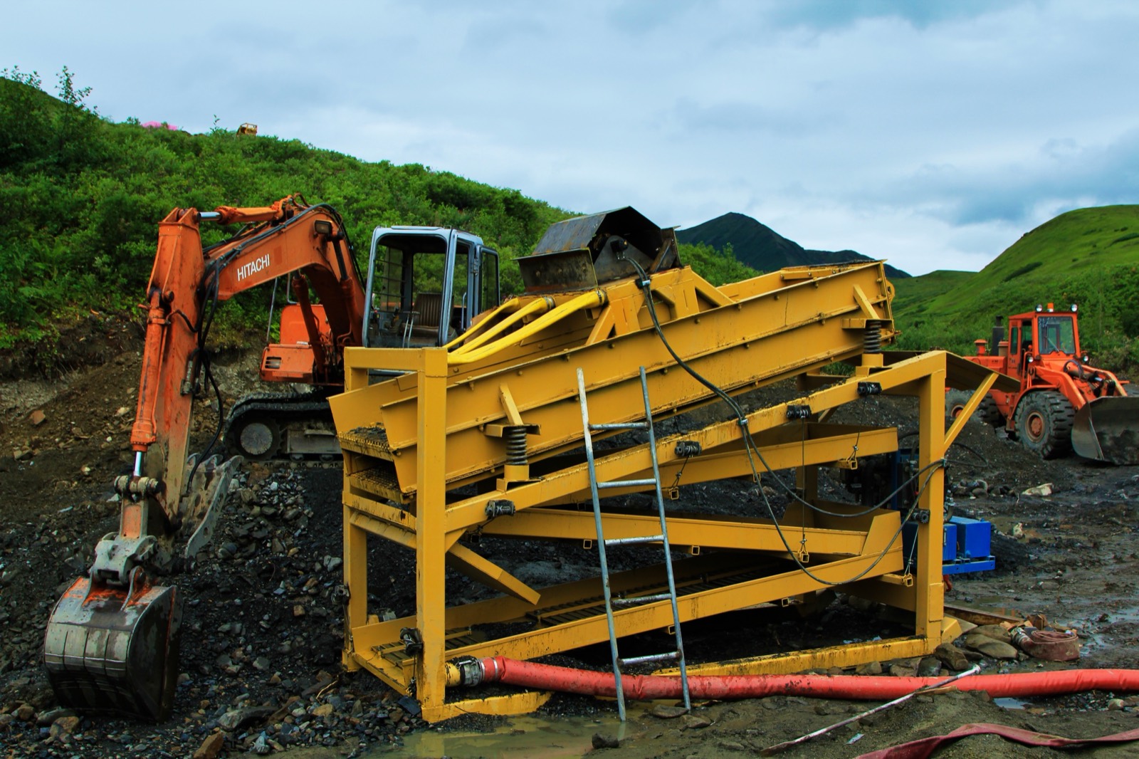 M100 gold wash plant on a placer claim, excavator feeding pay dirt into the hopper, tundra hills behind
